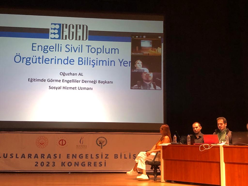 Fotoğrafta, yeşil ceket giymiş bir halde bir masada oturuyorum. önümde mikrofon ve fizüstü bilgisayarım bulunuyor. sunum ekranında adım ve sununun başlığı yazıyor.