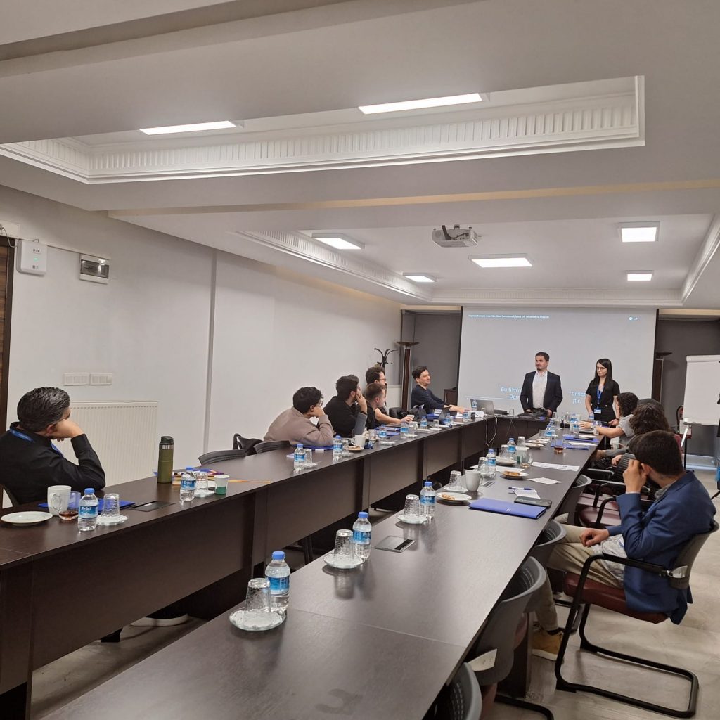 Fotoğrafta, modern bir toplantı odası görülüyor. Oda, uzun bir toplantı masası ve her biri önünde su şişeleri olan siyah ofis sandalyeleri ile döşenmiş. Masanın etrafında oturan yaklaşık on kişi var. Masanın bir ucunda, Oğuzhan AL ayakta duruyor ve bir projeksiyon ekranına doğru konuşuyor gibi görünüyor. Tavan, beyaz renkte ve yerleşik ışıklarla aydınlatılmış. Odanın bir tarafında beyaz bir duvar var ve diğer tarafta bir cam duvar var. Masanın üzerinde not defterleri, kalem ve belgeler var.